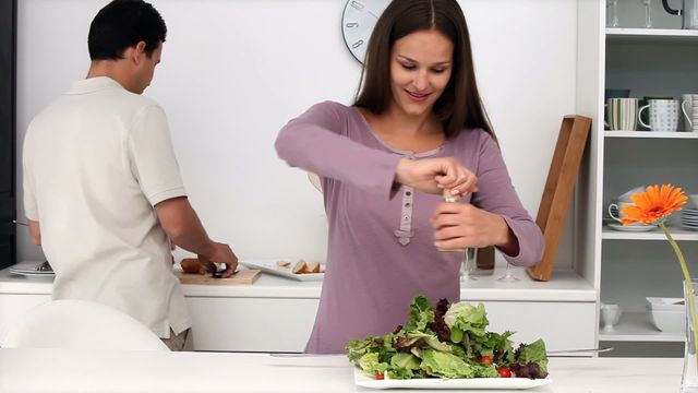 Couple enjoying cooking a healthy meal together in their modern kitchen. The woman is seasoning a fresh salad while the man slices bread in the background. Perfect for concepts of healthy living, relationships, teamwork, and modern domestic life.