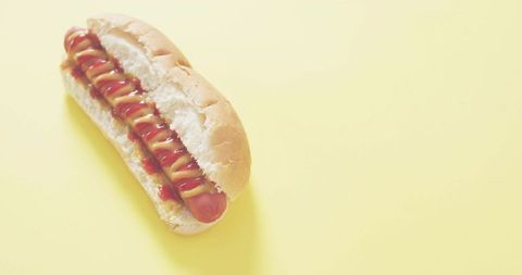 Sitting hot dog featuring mustard and ketchup drizzles on soft bun over yellow backdrop