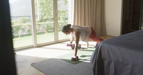 Woman exercising with dumbbells on yoga mat in sunlit bedroom for home fitness routine