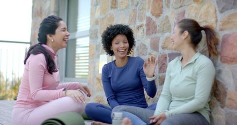 Diverse women laughing and chatting after yoga on sunlit wooden deck in pastel activewear