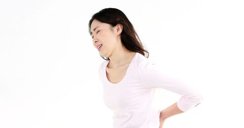 a young asian woman experiences stomach pain while standing against a white background, illustrating discomfort or health issues related to stomachache or back pain. useful for articles or advertisements focusing on health, medical advice, or wellness programs.