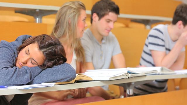 Shows female student sleeping during lecture while others study. Useful for representing academic exhaustion, exam stress, or group collaboration in educational settings. Represents college life, student challenges, and concentration in academics.