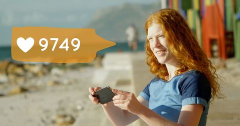Caucasian Redheaded Woman Texting at Beach with Social Media Icon