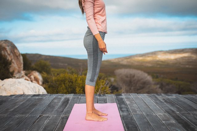 Caucasian woman standing on a pink yoga mat on a wooden deck in a mountainous area. Ideal for promoting outdoor fitness, healthy lifestyle, yoga retreats, meditation practices, and travel adventures.