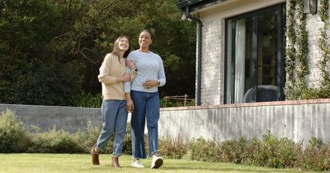 Female couple walking arm-in-arm on lawn by home, sharing garden stroll and companionship