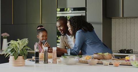 African american family preparing salad on modern kitchen island, smiling and sharing smartphone
