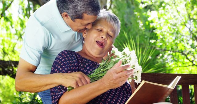 A senior Asian couple shares a tender moment outdoors, with the man kissing the woman's cheek as she holds a bouquet and a book, with copy space. Their affectionate gesture and relaxed environment suggest a comfortable and loving relationship.