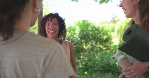 Diverse women chatting after outdoor yoga in leafy garden holding mat and bottle