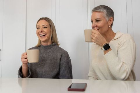 Two women sharing coffee and laughter at bright kitchen table, cozy sweaters and smartphone