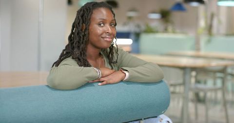 African woman leaning on teal bench, smiling and wearing smartwatch in coworking space