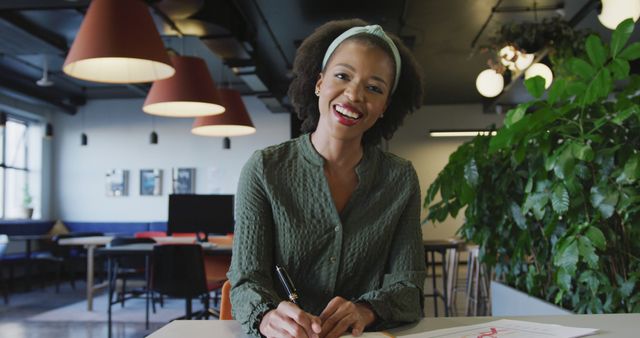 This image shows a smiling businesswoman seated at a modern office desk, working on documents. She appears to be engaged and cheerful, suggesting a positive work environment. Ideal for use in articles or websites discussing business, productivity, modern workplaces, office life, or employee satisfaction.