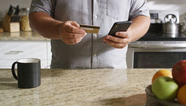 Man making mobile contactless payment at home kitchen counter with credit card smartphone