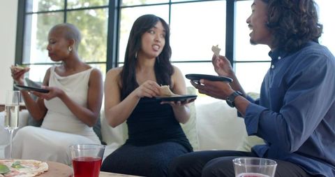 Friends sharing pizza on couch during casual indoor gathering with drinks and conversation