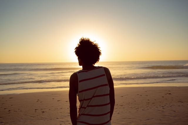 This image captures a man standing on the beach at sunset, looking out at the ocean. The scene evokes feelings of contemplation and tranquility, making it ideal for use in travel brochures, wellness blogs, inspirational content, and advertisements promoting relaxation and peaceful retreats.