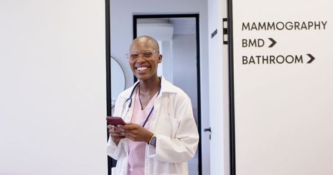 Smiling clinician holding smartphone at clinic door, wearing lab coat, scrubs and stethoscope