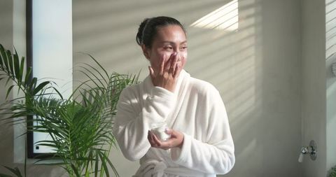 Asian woman applying moisturizer in sunlit minimalist bathroom during morning skincare ritual
