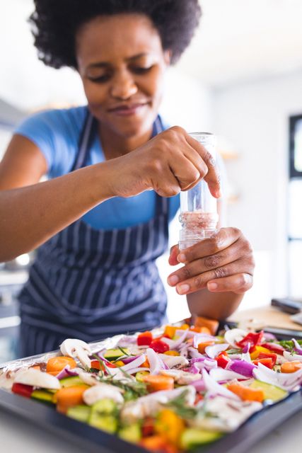 African american mid adult woman sprinkling seasoning on food in kitchen. unaltered, lifestyle, preparation, cooking, food, vegetable.