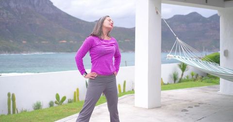 Middle-aged woman stretching on coastal veranda near hammock, seascape wellness retreat
