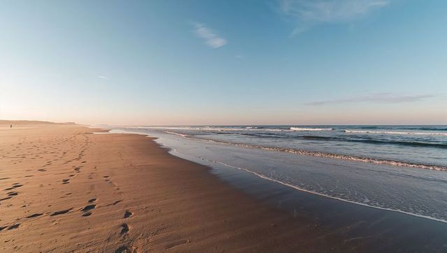 Wet sand reflecting sky on empty beach at low tide with footprints and lone walker at dawn