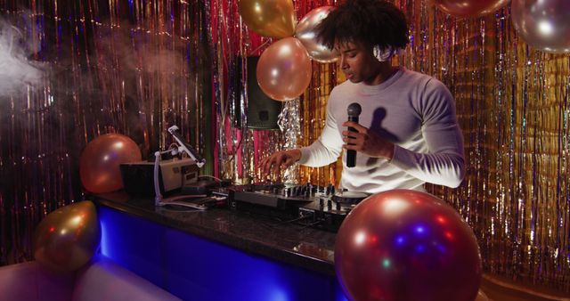 Image of happy african american male dj playing music, dancing and using microphone at a nightclub. Fun, inclusivity, going out and party concept.