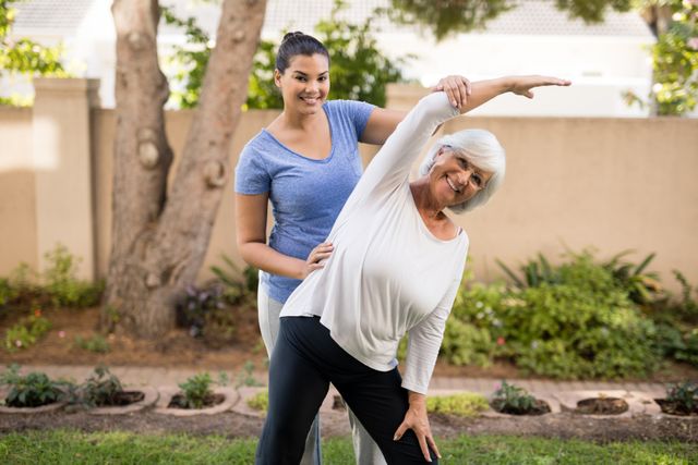 Senior woman receiving assistance from a trainer while stretching in a park. Ideal for promoting fitness programs for seniors, personal training services, healthy lifestyle campaigns, and active aging initiatives.