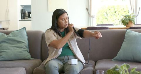 Asian woman checking blood pressure at home on sofa using digital cuff monitor