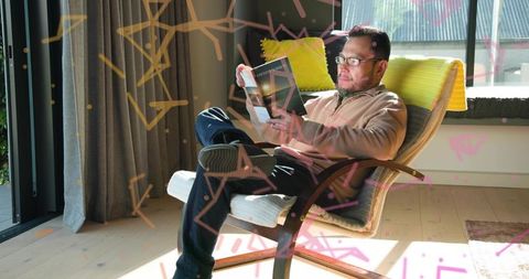Serene asian man relaxing and reading glossy magazine on sunlit lounge chair at home