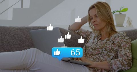 Reclining woman using laptop receiving social likes on grey sofa in bright living room