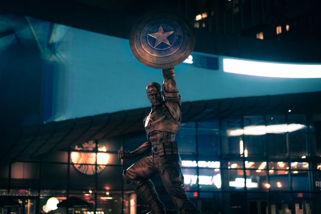 Captain America statue holding shield aloft at night in urban environment, well-lit with buildings in background. Iconic for superhero enthusiasts, perfect for illustrations of nocturnal city scenes, or superhero-themed promotions.