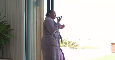 African american woman enjoying morning tea while using smartphone by sunlit sliding door