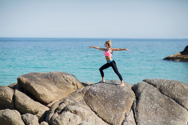 Woman practicing warrior pose on rocky shore with ocean in background. Ideal for promoting fitness, yoga, outdoor activities, and healthy lifestyle. Suitable for use in wellness blogs, fitness magazines, and travel brochures.