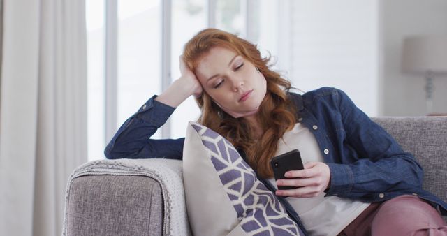 Young redheaded woman sitting on couch, looking at her smartphone with bored expression. This can be used for themes related to boredom, social media, technology addiction, modern lifestyle, or relaxation at home.