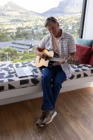 Senior man sitting on window seat playing acoustic guitar with laptop and mountain view