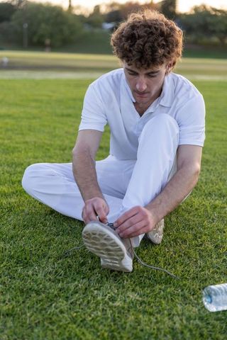 Young man tying shoelace on grassy field wearing white polo and trousers at golden hour