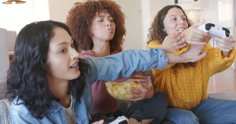 Multicultural Friends Playing Video Games and Sharing Snacks on Couch in Cozy Living Room