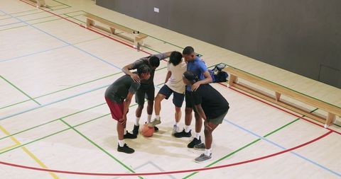 Diverse male friends huddling on indoor court, dribbling basketball and planning team play