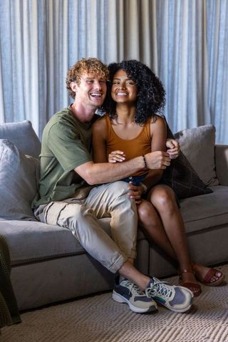 Smiling diverse couple cuddling on light-gray sofa in cozy living room with cushions and rug