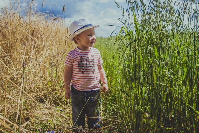 This image features a joyful toddler wearing a straw hat, standing in a green field with tall grass on a summer day. The child is dressed in a striped shirt and jeans, smiling and enjoying the natural surroundings. This photo can be used for diverse content related to childhood, outdoor activities, family life, carefree moments, and nature's beauty.