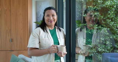African american woman smiling holding coffee mug by glass door with garden reflection