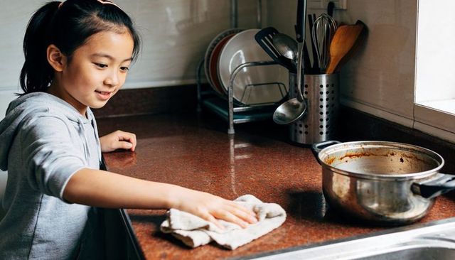 Asian girl wiping kitchen countertop with cloth, learning chores, smiling, daylight window