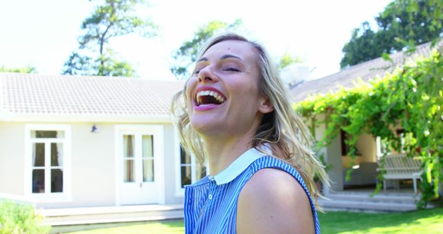 Woman laughing joyfully in a sunny outdoor setting with modern house in background. Suitable for concepts of happiness, joy, summer lifestyle, carefree living, real estate advertisements, and outdoor activities.