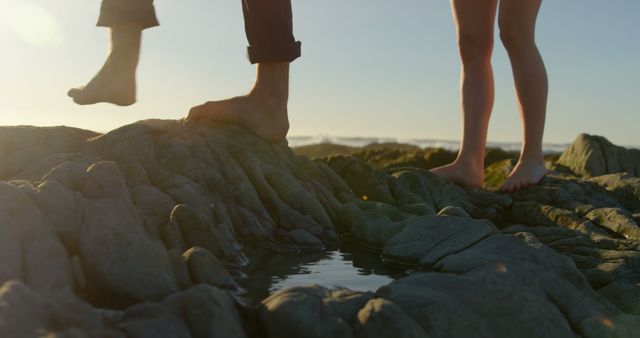 Couple standing barefoot on rocky shore during sunset. Useful in content focusing on outdoor activities, romance, travel, relaxation, or natural settings. Suitable for promoting beach destinations, romantic getaways, or lifestyle blogs.