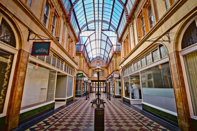 Image shows an empty historic indoor shopping arcade with a glass ceiling. The corridor features vintage architecture, including ornate tiles, large windows, and a glass canopy. Good for use in articles about historical shopping locations, urban architecture, revitalized shopping centers, and heritage tourism.