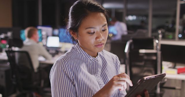 Depicts an Asian businesswoman using a digital tablet in a modern office setting at night. Ideal for content on technology in business, late-night work culture, professional settings, and digital communication. Can be used for articles, blog posts, marketing materials related to office productivity, technology use, and corporate environments.