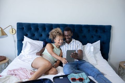 Mixed-race couple relaxing on bed sharing tablet while packing for weekend getaway