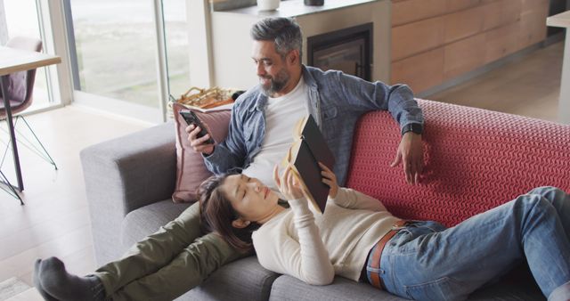 Couple is relaxing on couch in modern living room, man using smartphone and woman reading book. Ideal for depicting home life, leisure activities, technology use, relaxation, and modern home interiors.