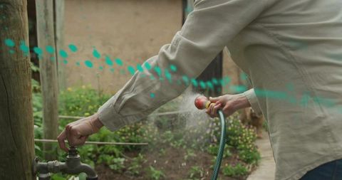 Mature woman watering backyard garden turning outdoor spigot with hose spray among flowers