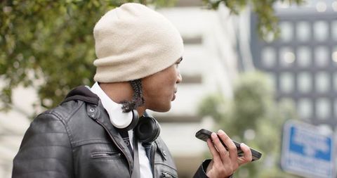 Young man speaking into smartphone on city sidewalk wearing beanie and headphones