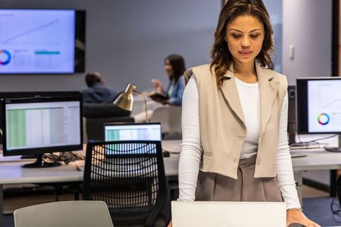 Woman in 30s standing at desk using laptop in modern open office with charts on screens