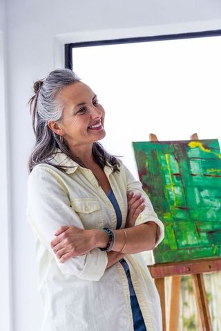 Senior woman artist standing in bright studio smiling beside vibrant abstract green canvas